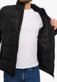 Chaqueta acolchada negra con cierre frontal, con un diseño cuadrado acolchado, cuello alto y un forro interior suave. Lleva puesta sobre una camiseta blanca.