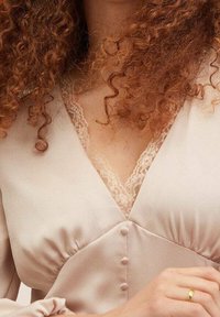 Des boucles rouges tombent sur un blouse en satin crème avec des bordures en dentelle et des boutons recouverts de tissu, la main reposant près de la taille avec une bague en or.