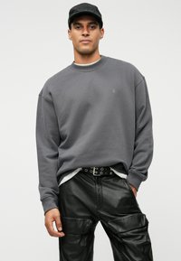 Άνδρας που φοράει ένα σκούρο γκρι φούτερ, μαύρο καπέλο, μαύρο δερμάτινο παντελόνι cargo και μια μαύρη ζώνη με ασημένια κουμπώματα σε απλό φόντο.