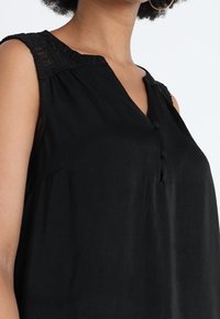 Musta ilmanhiha toppi, jossa on V-pääntie, pitsiyksityiskohtia olkapäillä, napitus edessä ja löysä istuvuus, valmistettu kevyestä kankaasta.