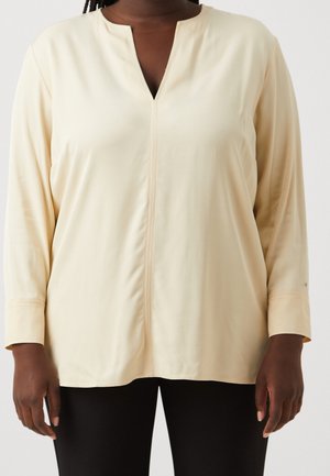 Kvinde iført en cremefarvet langærmet bluse med V-slids ved halsudskæringen og sorte bukser, stående foran en ensfarvet baggrund.