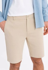 Pantaloni beige con tessuto a trama, chiusura frontale con bottone e tasche laterali, abbinati a una camicia blu chiaro e a una maglietta bianca.