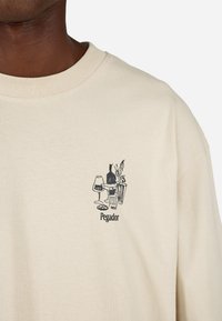 Bežs kokvilnas T-kreklu ar apaļu kaklu, uz kura ir melns zīmējums ar brillēm, pudeli un augu, un zem tā ir teksts "Pegador".