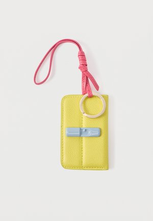 Geel leren sleutelhangerzakje met roze polsbandje, gouden sleutelring en blauw, penvormig sierstuk op witte achtergrond.