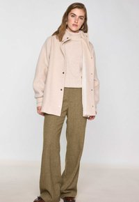 Giacca in misto lana beige con colletto alto e bottoni a pressione, abbinata a un dolcevita beige chiaro e pantaloni a gamba larga verde oliva strutturati.