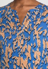 Haut à motifs bleus présentant un design abstrait en beige et noir. Le tissu a une texture lisse avec un col en V et des manches courtes.