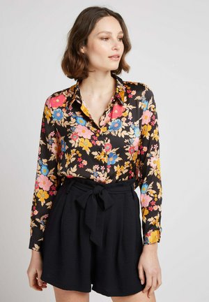 Blouse noire à imprimé floral avec des fleurs multicolores en rose, bleu et jaune. Manches longues, détail de col, associée à un short noir taille haute.