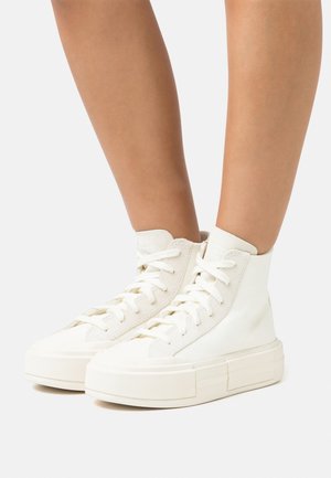 White high-top sneakers feature a smooth textile upper, thick rubber soles, and flat laces. The design includes a subtle seam detail.