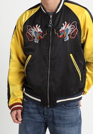 Veste bomber noire et jaune avec broderie de dragon sur la poitrine, poignets et ceinture rayés, portée avec un jean bleu.