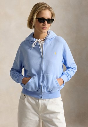 Kobieta w czarnych okularach przeciwsłonecznych, jasnoniebieskiej bluzie z kapturem na zamek z rękami w przednich kieszeniach i białych spodniach na neutralnym tle.