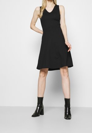 Kobieta w czarnej, bezrękawnikowej sukience do kolan i czarnych botkach na obcasie, stojąca z jedną ręką na biodrze na białym tle.