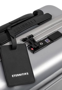 Silberner Hartschalensuitcase mit matter Oberfläche, schwarzem Griff und einem Gepäckanhänger. Verfügt über ein Schloss und ein Kombinationsschloss an der Seite.