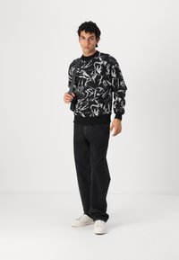 Black sweatshirt featuring a bold white abstract pattern, paired with black jeans and white sneakers. Model holds a gray shoulder bag.