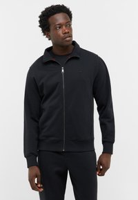 Felpa nera con zip realizzata in tessuto liscio, con colletto alto, polsini a coste e un logo semplice sul petto.