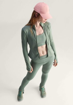 Sportovní set v tlumené zelené barvě obsahuje bundu na zip, crop top a legíny, doplněné růžovou čepicí a šedými teniskami. Zvýrazňují ho texturované detaily.