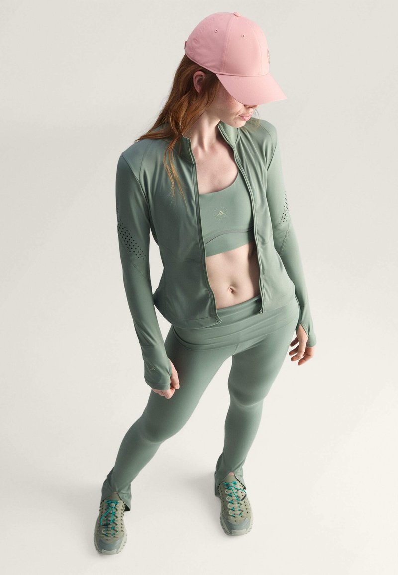 Athletic set in muted green includes a zip-up jacket, crop top, and leggings, complemented by a pink cap and grey sneakers. Features include textured accents.