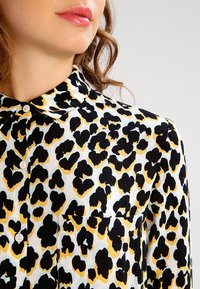 Blusa de leopardo con fondo blanco, patrones en negro y amarillo, cuello estructurado y cierre frontal con botones, además de un bolsillo en el pecho.