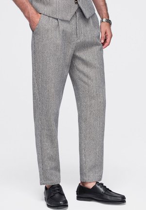 Homme portant un pantalon de costume gris à motifs, des chaussures en cuir noires et un gilet assorti, les mains dans les poches.