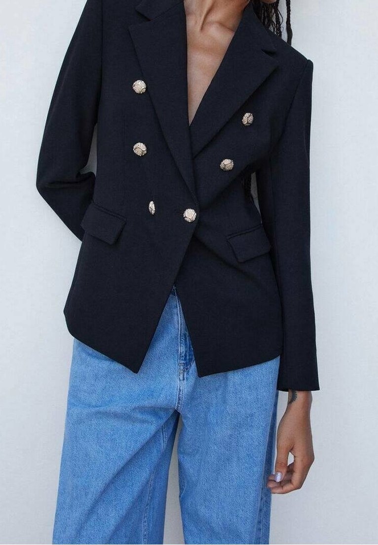Personne portant un blazer noir croisé avec des boutons dorés et un jean taille haute bleu clair sur un fond uni.