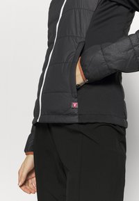 Personne portant une veste matelassée noire, une main dans la poche latérale, une fermeture éclair blanche visible, et un pantalon noir sur un fond uni.