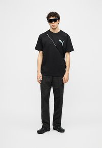 Νεαρός άνδρας φορώντας μαύρο μπλουζάκι Puma, μαύρο παντελόνι, μαύρα παπούτσια, σκουρόχρωμα μεγάλα γυαλιά ηλίου και τσάντα με ιμάντα χιαστί σε λευκό φόντο.