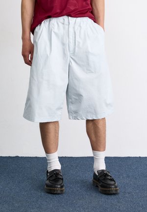 Personne portant un short blanc ample jusqu'aux genoux, des chaussettes blanches, des mocassins noirs à pompons et une chemise rouge, les mains dans les poches.