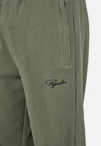 Olivové kordové nohavice s vertikálnou rib iba textúrou, s bočnými vreckami a čiernym vyšitým logom "Pegadon" na stehne.