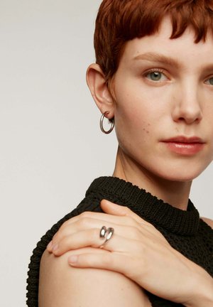 Pendiente de aro plateado y un anillo curvado de plata, llevados por una persona con cabello corto y rojo, vestida con un suéter negro de punto texturizado.