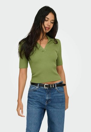 Kvinne med langt mørkt hår iført en grønn, ribbestrikket kortermet topp og blå jeans med svart belte, stående mot en ensfarget bakgrunn.