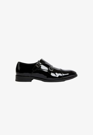 Zapato de charol negro con acentos de doble hebilla, punta redondeada y tacón bajo. Textura lisa y brillante con una silueta elegante.