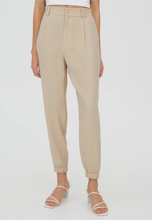 Pantalons beiges taille haute à coupe décontractée, avec des bas resserrés, des plis devant et une fermeture à un bouton, fabriqués en tissu lisse.