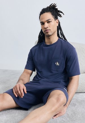 Άνδρας με μακριά ντραντλόκ, φοράει μπλε σκούρο t-shirt Calvin Klein και σορτς, κάθεται σε ανοιχτό γκρι χαλί με μαξιλάρι.