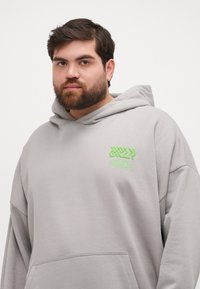 Hanorac supradimensionat de un gri deschis, cu un logo brodat verde pe piept și o buzunar tip cangur. Material moale, cu o croială lejeră și glugă.