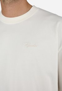 Cămașă din bumbac alb cu mâneci lungi și guler rotund. Prezintă un logo brodat mic, subtil, de culoare bej pe piept. Textură fină.
