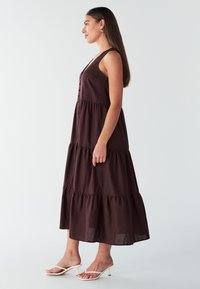 Mørkebrun bomulds maxi-kjole med lagdelt design, v-udskæring, knapper foran og tynde stropper. Kombineret med hvide sandaler.