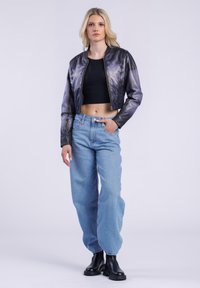 Ung kvinde står med hånden i lommen, iført en kort sort top, metalisk bomberjakke, løse lyseblå jeans og sorte støvler.