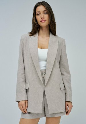 Jeune femme portant un blazer beige ajusté et un short assorti avec un haut blanc, debout devant un fond gris clair uni.