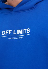 Felpa blu con testo bianco che legge "OFF LIMITS" sopra una linea e "DANGEROUS AREA" sotto, indossata da una persona.