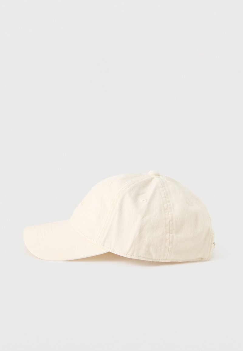 Gebroken witte katoenen baseballpet met gebogen klep en zichtbare stiksels, vanaf de zijkant getoond tegen een eenvoudige lichte achtergrond.