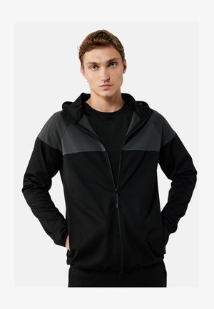 Jonge man met kort bruin haar, draagt een zwarte zip-up hoodie met donkergrijze panelen en handen in de zakken, kijkt recht vooruit tegen een witte achtergrond.