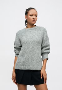 Grau melierter Wollmischpullover mit Rundhalsausschnitt, lässiger Passform und voluminösen Ärmeln, getragen über einem schwarzen plissierten Minirock.