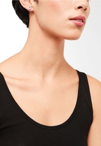 Schwarzes, geripptes Tanktop mit einem runden Ausschnitt, kombiniert mit einem kleinen, runden Ohrring. Die Haut wirkt glatt und hat natürliche Farbtöne.
