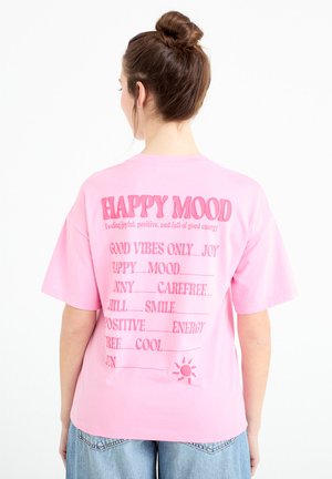 Ung kvinde med brunt hår sat op i en knold bærer en løs lyserød T-shirt med positive ord og en solgrafik på ryggen.