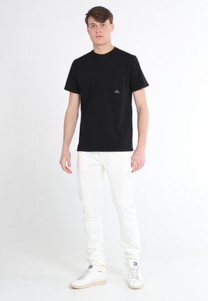 T-shirt nera in cotone con colletto rotondo e taschino sul petto, caratterizzata da un piccolo logo triangolare, abbinata a jeans bianchi slim-fit e sneaker.