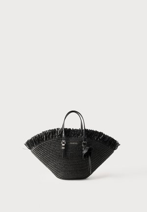 Bolso tote tejido negro con forma amplia de abanico, borde superior con flecos, dos asas y un lazo decorativo en una de las asas.