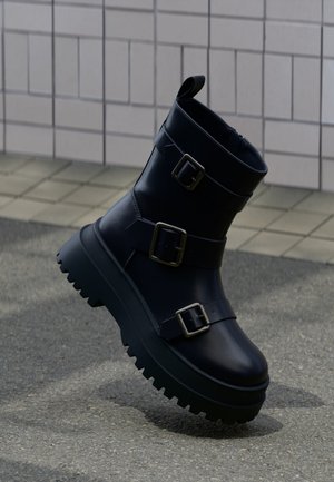 Schwarzer Lederknöchelstiefel mit klobiger Sohle und drei verstellbaren silbernen Schnallen, gezeigt auf einem gefliesten und asphaltierten Untergrund.