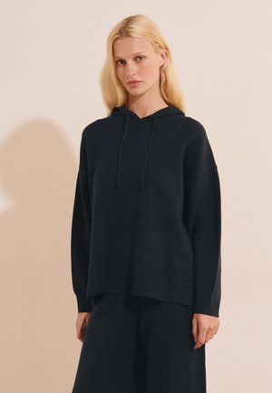 Femme blonde aux cheveux raides porte un pull à capuche noir ample et un pantalon noir assorti, debout devant un fond beige.