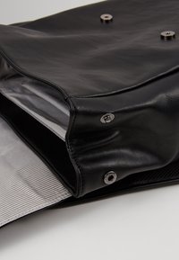 Sac en cuir noir avec fermetures à pression argentées, ouvert pour montrer la doublure intérieure en tissu rayé gris, exposé sur une surface blanche.