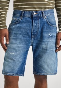 Pantalones cortos de mezclilla en azul claro con un acabado desgastado y detalles rasgados. Cuenta con cinco bolsillos y un cierre de botón en la cintura.