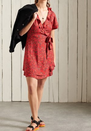 Vestido cruzado rojo con flores, mangas cortas con volantes, cintura con lazo y dobladillo en capas. Llevado con una chaqueta negra y sandalias negras con suelas naranjas.